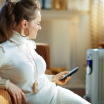 A woman sits against a sofa while using a remote to operate a portable room heater for supplemental apartment heating.